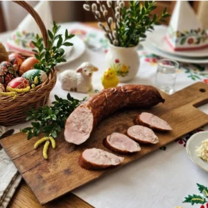 Tradycyjna kiełbasa koszyczkowa marki Wędlina z Lublina, serwowana na drewnianej desce na bogato zdobionym stole wielkanocnym z pisankami i baziami.